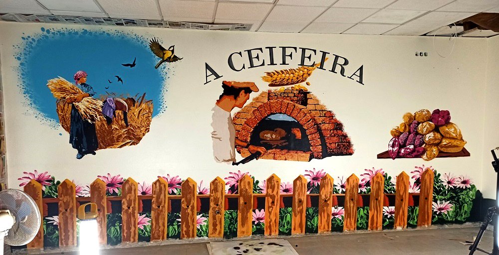 Os três murais do "Ciclo do Pão" (As Ceifeiras, O Forno e o Padeiro, A Celebração do Pão) juntos na Padaria A Ceifeira. Na base, uma cerca pintada com malmequeres cor de rosa une visualmente as três obras.