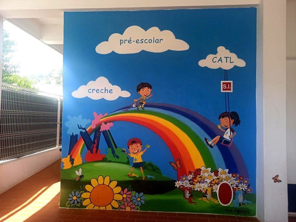 Mural "A Alegria de Pintar" na Casa do Povo de Messines. O Arco-Íris central liga as valências (Creche, Pré-escolar, CATL), com crianças alegres (manchadas de tinta) a brincar sobre ele e num campo de flores estilizadas.