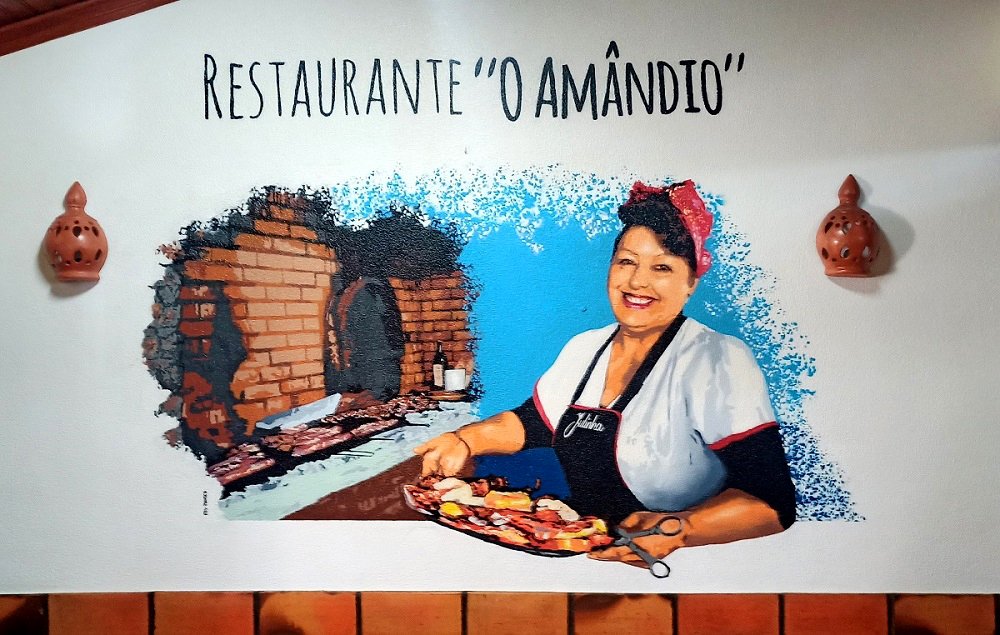 Mural no Restaurante O Amândio (V. N. Milfontes) com o retrato da Julinha, a anfitriã, sorridente, com lenço e avental. Ela apresenta a especialidade da casa (grelhados), celebrando a hospitalidade do espaço.