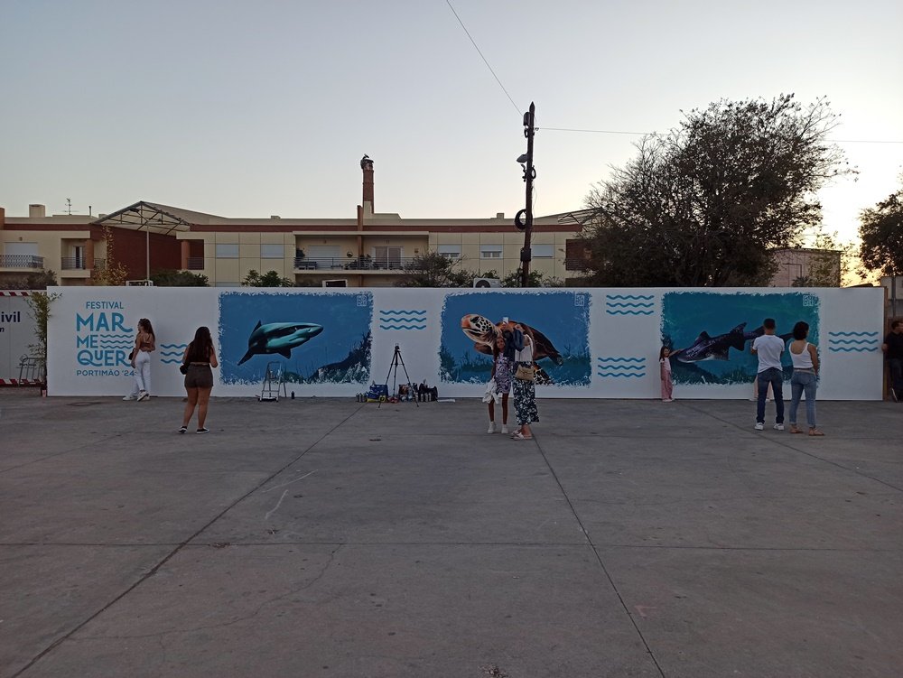 Mural terminado no Festival Mar Me Quer em Portimão. Vista de longe, com várias pessoas a tirar selfies a média distância para apanhar o mural todo. Outras pessoas estão junto ao mural a tirar fotos, interagindo com a arte urbana de conservação marinha.
