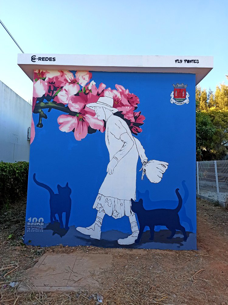 Mural "Homenagem à Marquinhas" em Almancil. A figura é representada em traço minimalista, rodeada por gatos em silhueta e flores de amendoeira, sobre um fundo azul. A obra eterniza a memória da querida figura da vila.