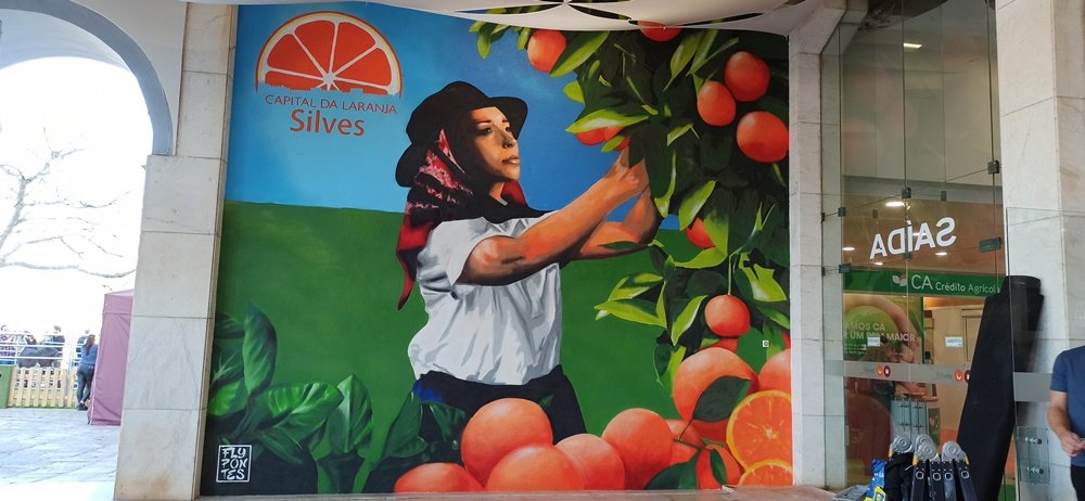 Mural "Mostra da Laranja 2025: Colheita Vibrante" terminado (5m×6m) em Silves. A obra celebra a tradição agrícola com uma jovem vibrante a colher laranjas contra um céu azul límpido.