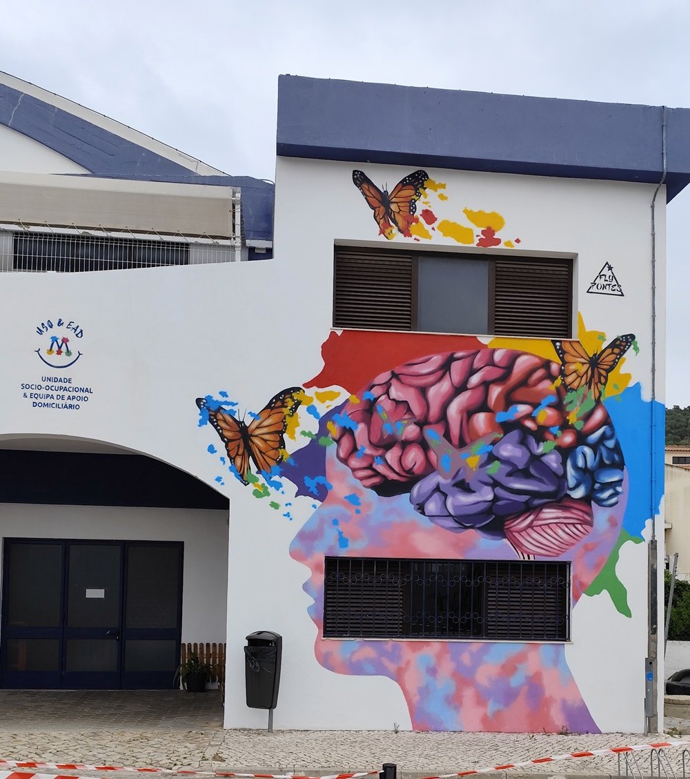 Mural "A Metamorfose" terminado, símbolo do trabalho da Unidade Sócio-Ocupacional (USO). A obra apresenta uma cabeça de perfil com o cérebro vibrante e três Borboletas-Monarca a emergir, representando a evolução e a criatividade.