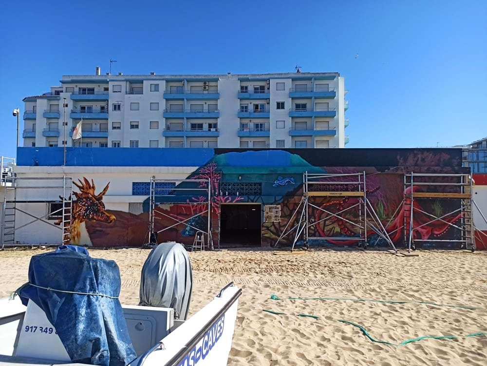 Vista remota do Mural Pedra do Valado, em Armação de Pêra, com pintura em progresso; todos os animais marinhos finalizados. A foto, tirada de um barco visível, mostra a escala da arte urbana na fachada.