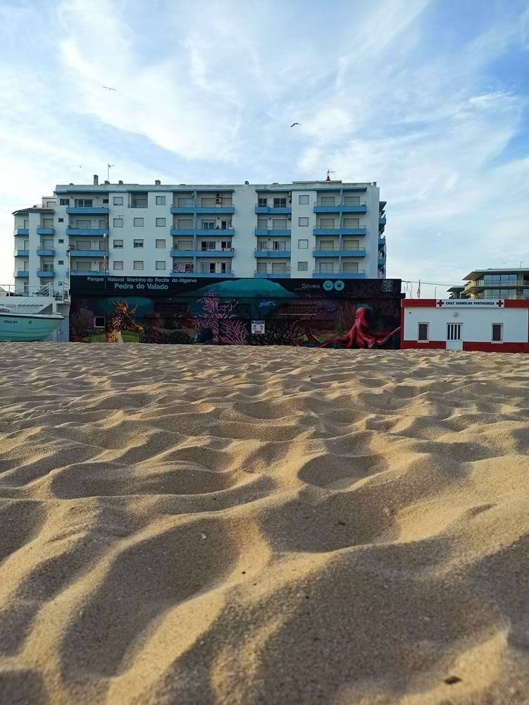 Mural Pedra do Valado totalmente terminado na Lota de Armação de Pêra, visto da areia. A foto enquadra o mural ao lado da Casa do ISN e os prédios da cidade, sob um céu azul com nuvens. Obra de arte urbana no Algarve.