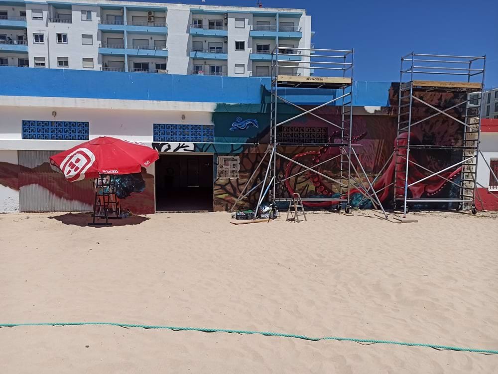 Vista geral da Lota de Armação de Pêra com portão aberto, mostrando a fase final da pintura mural Pedra do Valado. Visíveis os andaimes, o polvo, a lesma-do-mar, as gorgónias, um chapéu de sol e a cadeira junto à área do Mero. Arte urbana Algarve.