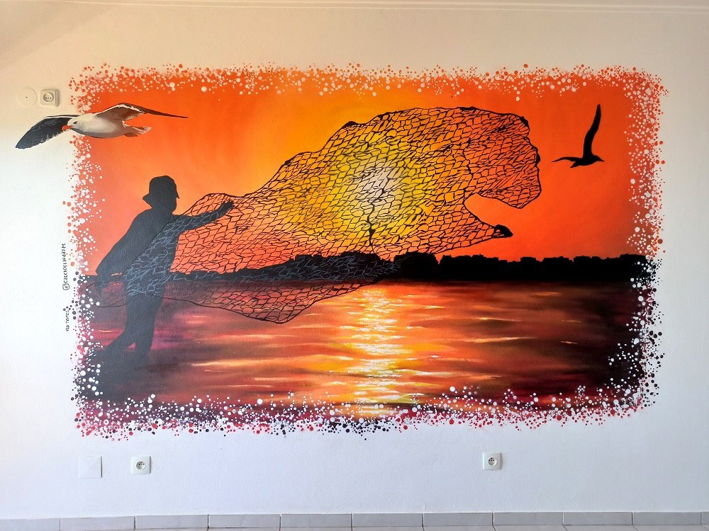 Mural "O Pôr do Sol do Pescador" (stencil e spray) criado para um Alojamento Local em Portimão. A silhueta do pescador a lançar a rede e a silhueta da cidade enquadram-se num pôr do sol algarvio em tons quentes de laranja.