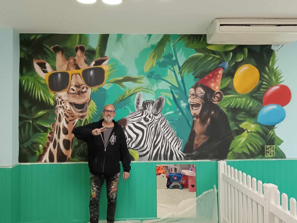 O artista Fly Pontes no meio do Mural da Selva (4x2,8m), com a pintura já pronta. O mural foi criado com spray à base de água. Imagem mostra a interacção do artista com a sua arte urbana vibrante no "Eat, Learn and Play" em Albufeira.