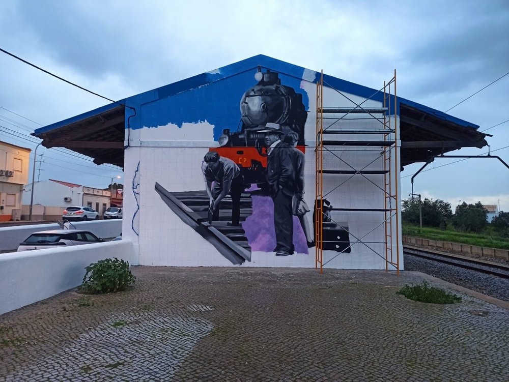 Parte central do mural "A Memória da Estação de Comboios" (Estação Almancil - Neche, Esteval) concluída. Vê-se a locomotiva a médio plano. Em primeiro plano, um homem de fato, gravata e chapéu, em tons de cinza, à espera do comboio, e uma mulher com malas de viagem. À esquerda, um trabalhador a construir o caminho de ferro. Andaimes montados à direita para a próxima fase da pintura mural.