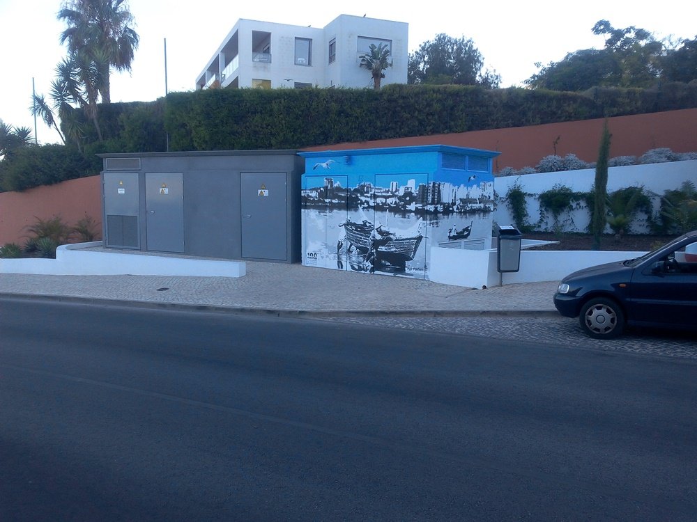 Mural "Homenagem aos Pescadores" concluído no posto de transformação (PT), visto do outro lado da estrada. A obra de dupla vista em tons de cinza e azul integra-se na paisagem de Portimão/Ferragudo, celebrando a tradição piscatória.
