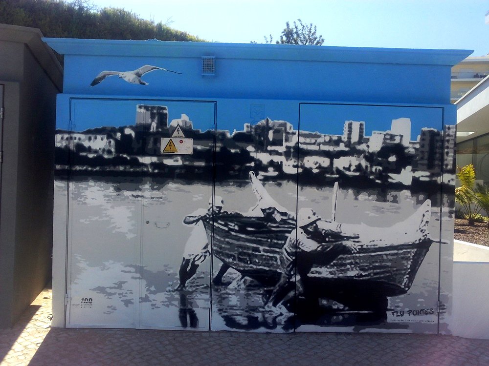 Vista frontal do mural "Homenagem aos Pescadores". Pescadores puxam o barco para terra na margem de Ferragudo, com a paisagem de Portimão visível ao fundo, simbolizando o trabalho árduo e a tradição piscatória.