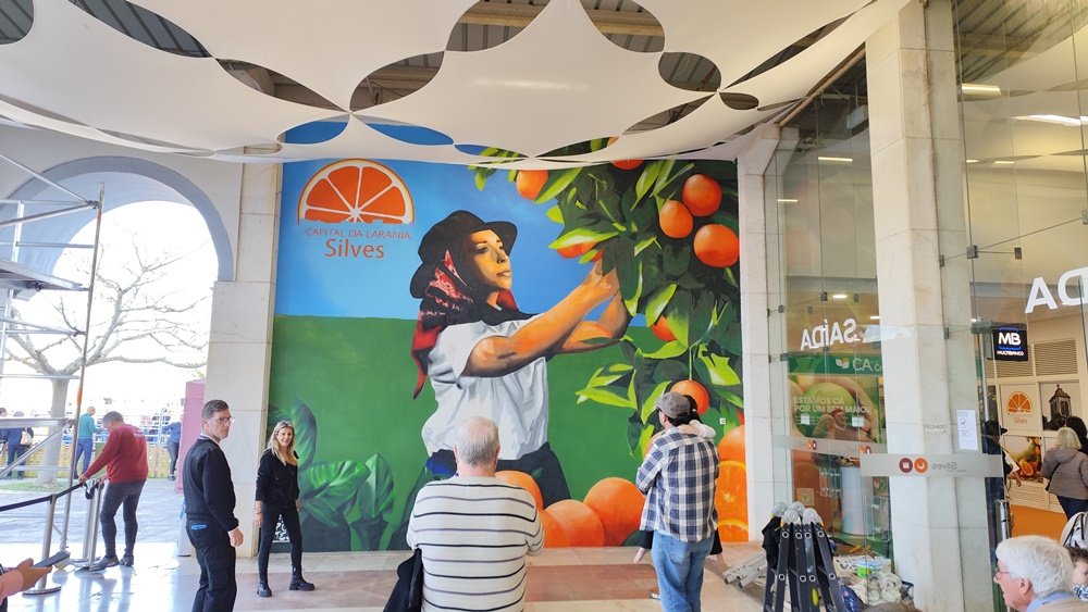 Mural "Mostra da Laranja 2025" concluído, com o público a apreciar a obra. Pessoas observam a pintura (5m×6m) em Silves, destacando o sucesso da arte urbana e a celebração da tradição agrícola.