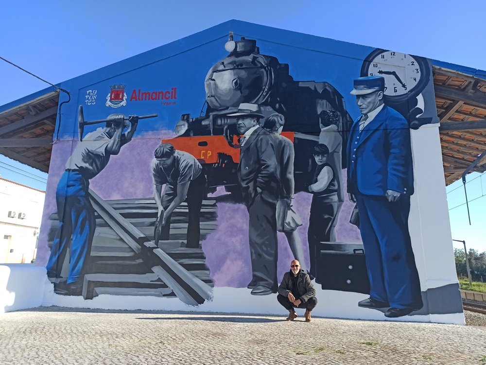 Mural totalmente terminado "A Memória da Estação de Comboios" na Estação Almancil - Neche (Esteval). Vista ao longe do mural de 12x8m com o artista Fly Pontes na imagem para dar perspectiva da dimensão. Arte urbana de nostalgia ferroviária.