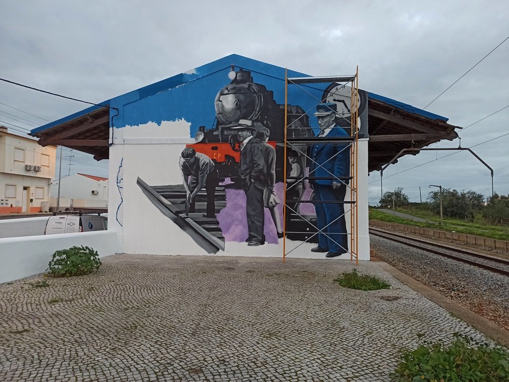 Detalhe do mural da Estação Almancil - Neche (Esteval). A parte direita e central mostra o Chefe da Estação vestido de azul, o relógio típico das estações portuguesas e as pessoas à espera do comboio (homem de fato, mulher com malas). Foco na recriação histórica da nostalgia ferroviária.