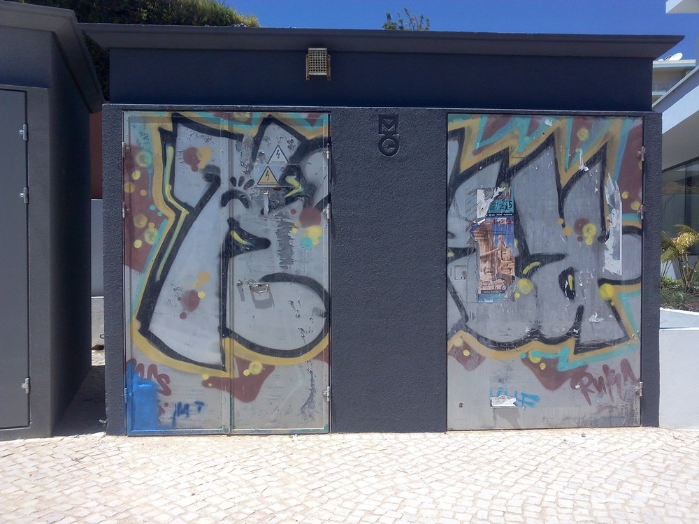 Foto do posto de transformação (PT) antes da pintura, com as portas cheias de graffiti. A estrutura funcional, em Portimão/Ferragudo, pronta para receber o mural "Homenagem aos Pescadores".