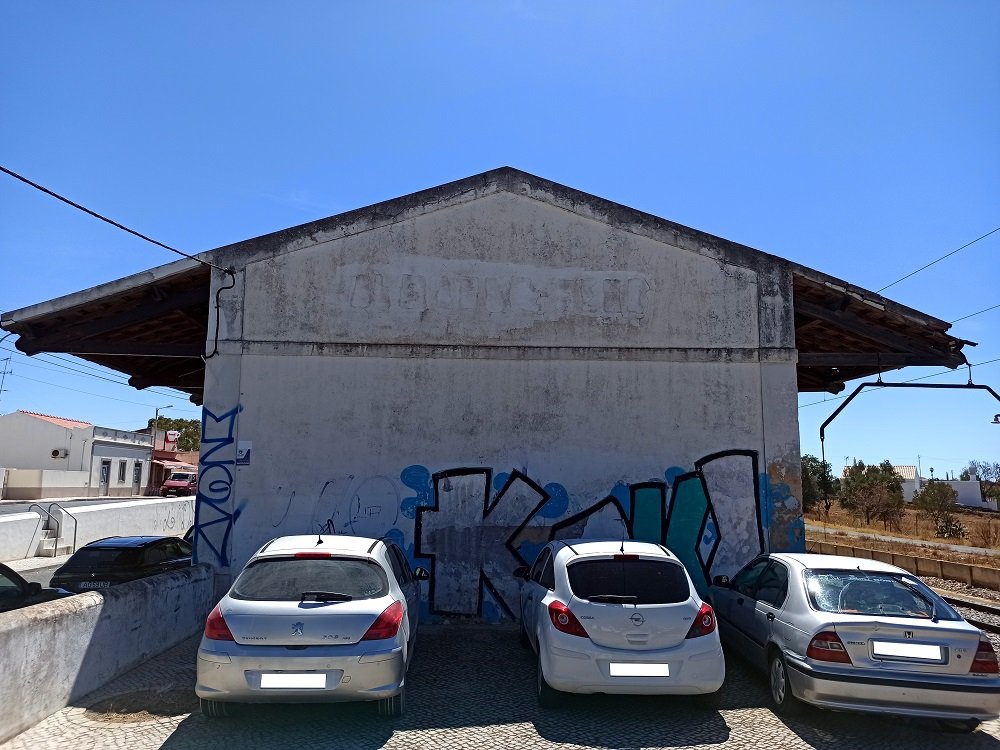 Foto de antes do projecto na estação Almancil - Neche (Esteval). Parede com grafites antigos e carros estacionados em frente. Vista do local antes da intervenção mural.