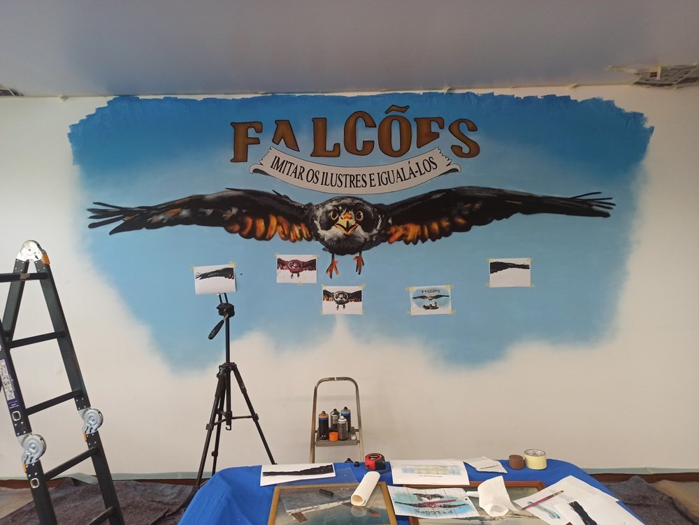 O Falcão e o lema "Imitar os Ilustres..." estão prontos no mural da BA11. Falta pintar o Castelo de Beja na parte inferior. Folhas de referência coladas na parede e latas de spray em cima do escadote, indicando o trabalho em curso.