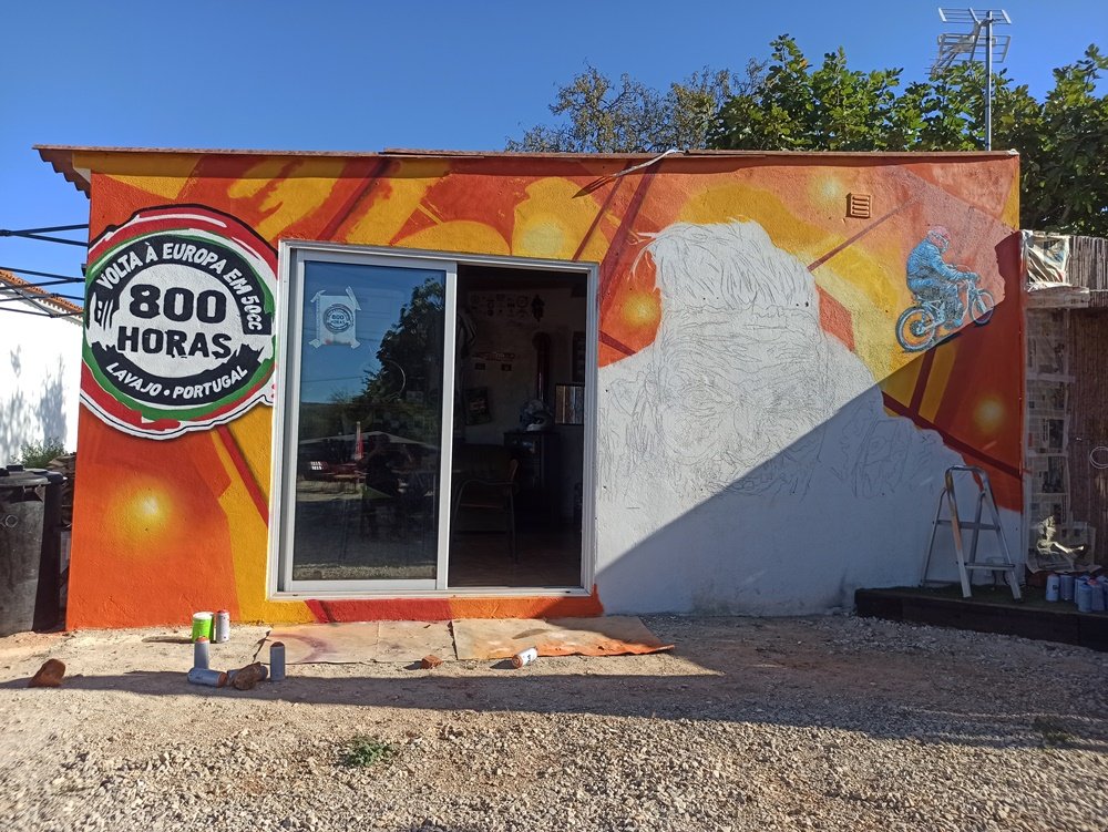 Fundo laranja, logotipo VEOH e motorizada terminados no mural "O Espírito da Estrada". A figura principal (Nelson Francês) está ao centro, desenhada, mas ainda sem cor, à espera do retrato realista. Latas de tinta no chão.