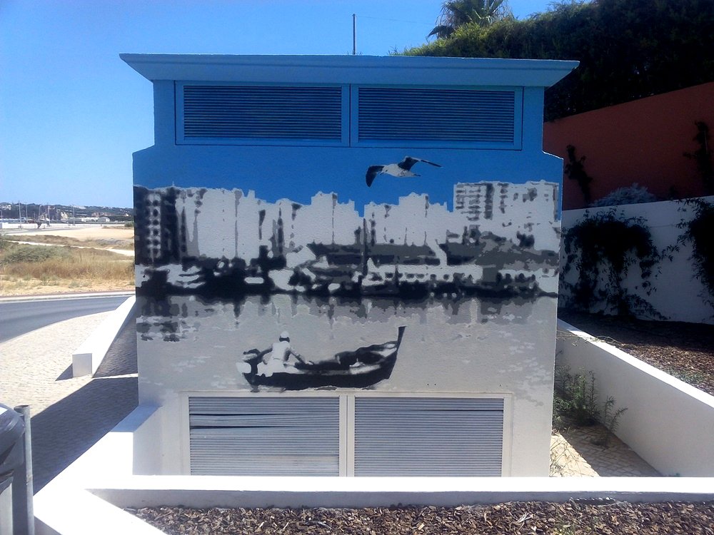 Vista lateral do mural "Homenagem aos Pescadores". Um pescador no barco a motor navega ativamente, visto da perspectiva de Ferragudo, em direcção ao skyline de Portimão ao fundo. Simboliza o regresso ao porto seguro no Algarve.