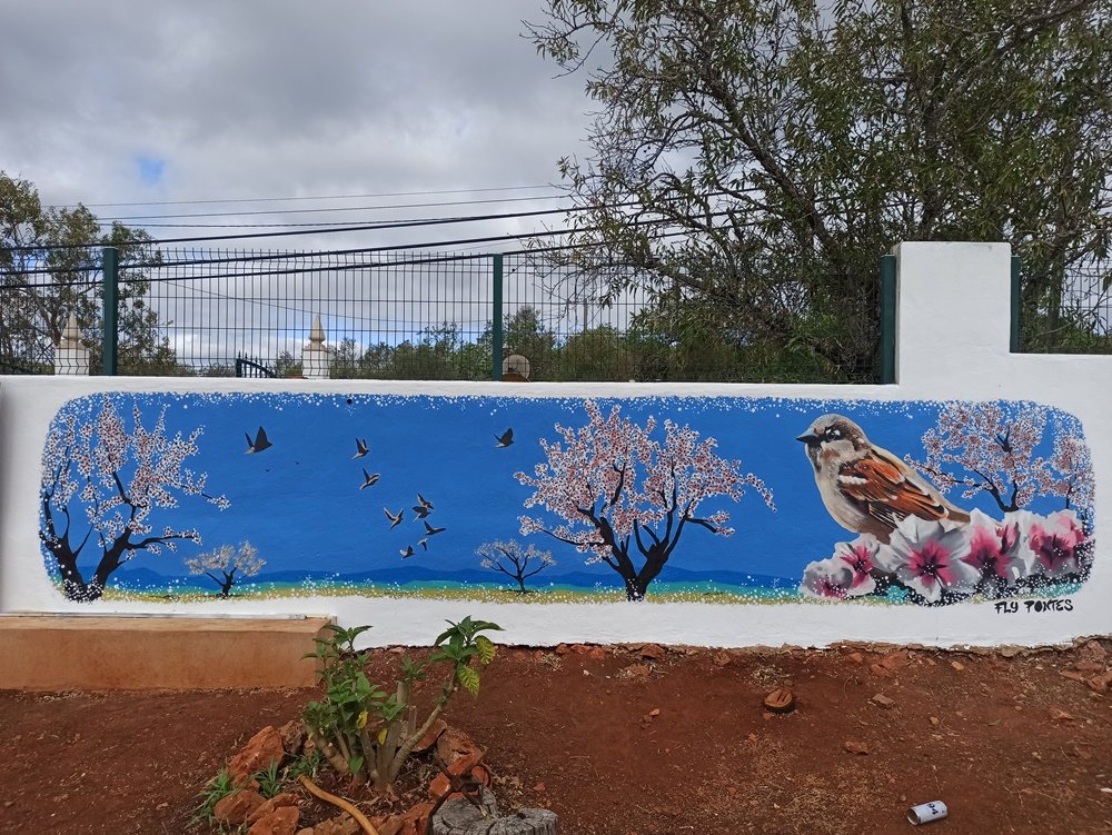 Mural "O Pardal no Despertar das Amendoeiras em Flor" (9m x 2,5m), inspirado no estilo japonês (Sakura). A pintura residencial celebra a Primavera algarvia com flores brancas e rosas e a presença do pardal, criando uma janela de tranquilidade e natureza.