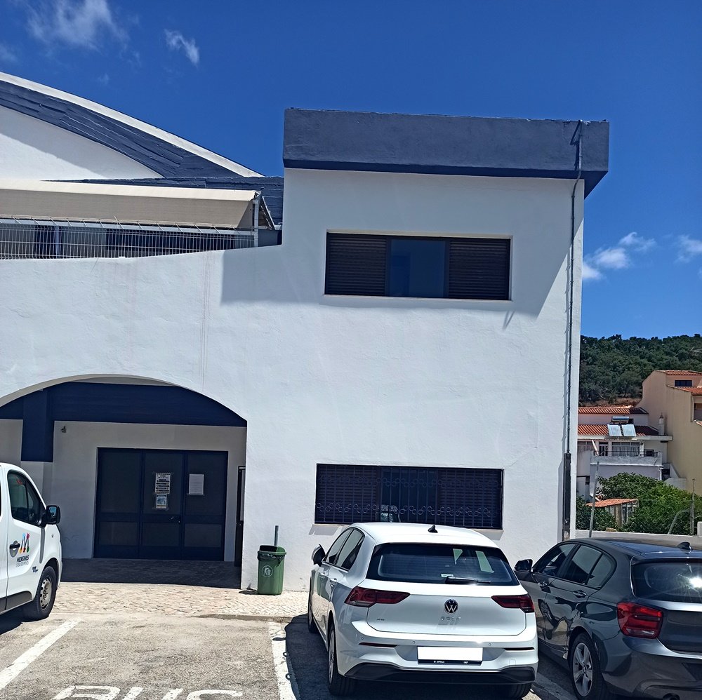 Foto de antes da intervenção do mural "A Metamorfose". Parede da Unidade Sócio-Ocupacional (USO) em branco, com alguns carros estacionados em frente. Vista do local antes da aplicação da arte urbana.