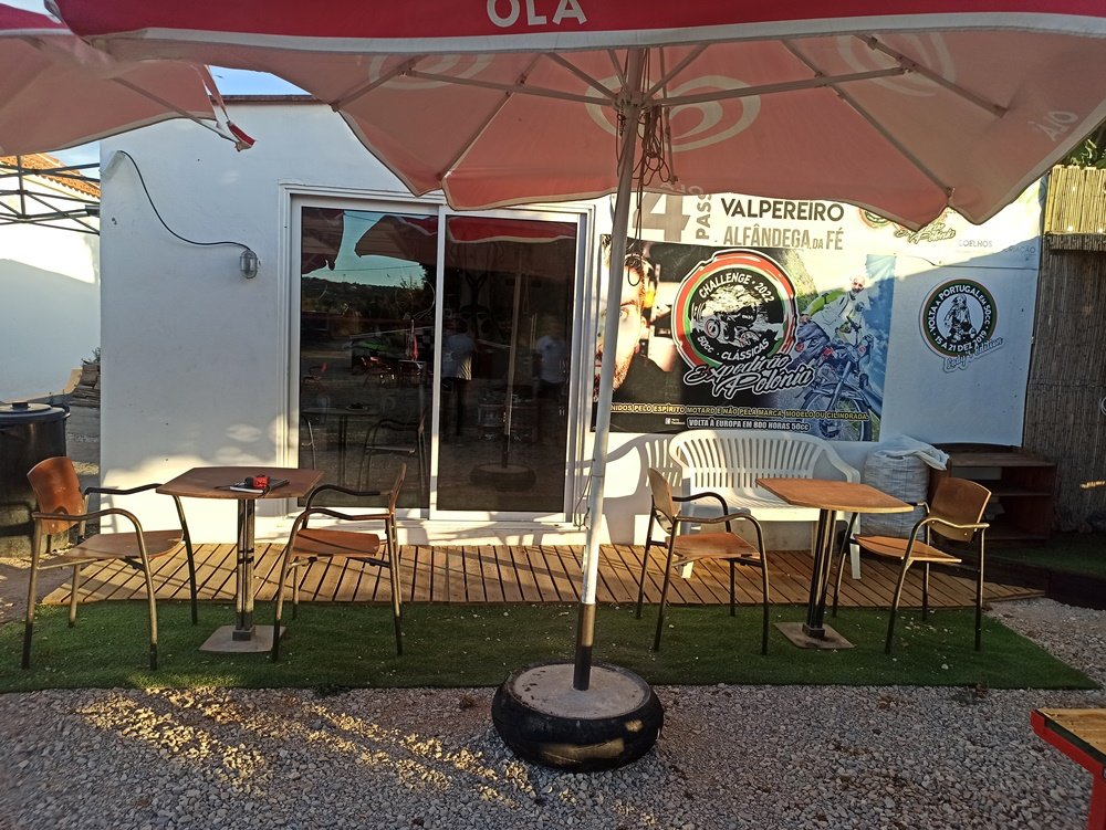 Foto da parede antes da pintura do mural da VEOH. A parede está cheia de cartazes de eventos anteriores e em frente veem-se mesas e cadeiras de esplanada. O local em Messines antes de receber o mural "O Espírito da Estrada".