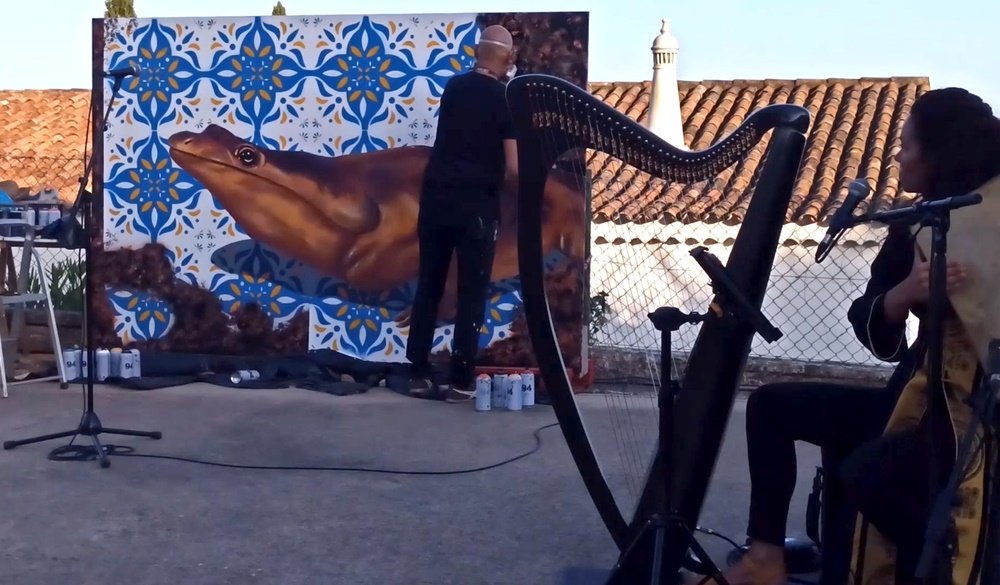 Performance ao vivo no GeoPalcos 2021: Helena Madeira a tocar adufe, com a harpa ao lado, em primeiro plano. Em segundo plano, o artista Fly Pontes a finalizar o mural, com uma chaminé algarvia ao fundo. A imagem capta a sinergia perfeita entre música e pintura.