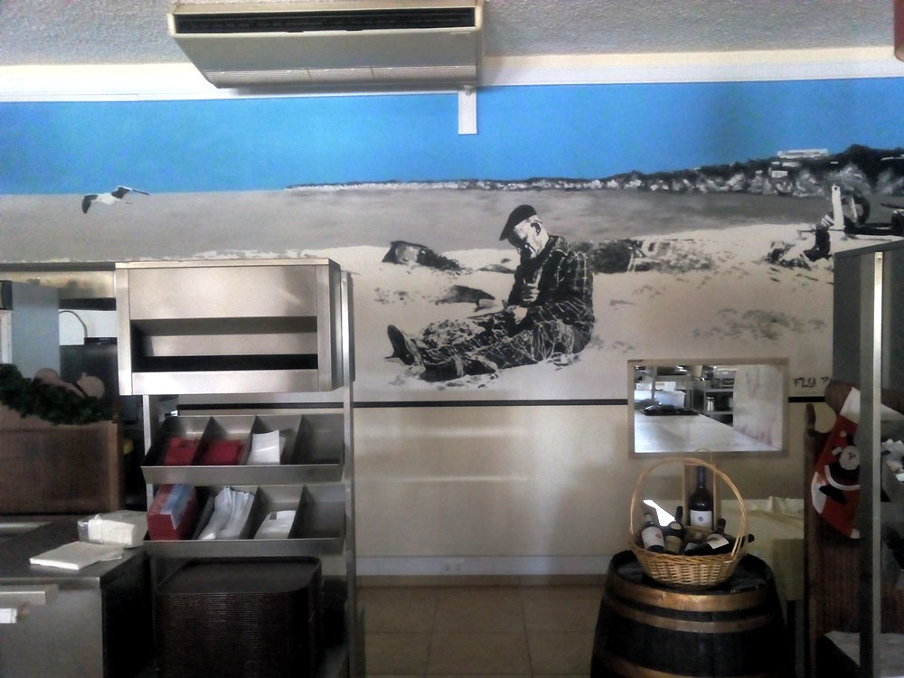 Vista do lado direito do mural de Armação de Pêra, com o Pescador a arranjar as redes e o Barco. A falésia em tons de cinza enquadra a cena, com a primeira Gaivota a voar no céu azul vibrante.