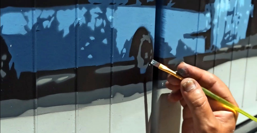 Plano aproximado da mão do artista Fly Pontes a segurar um pincel fino e a pintar um pormenor na pintura do autocarro. O pincel é usado para os retoques e acabamentos finais que garantem a definição dos detalhes.