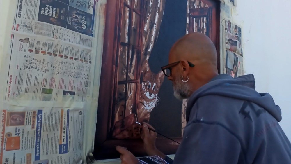 O artista Fly Pontes a aplicar os últimos retoques na moldura azul da janela com um pincel fino. O gato está visível no mural, com o seu olhar fixo no artista, reforçando a ilusão de óptica na fase final.