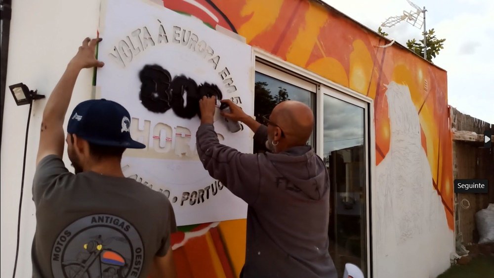 O artista Fly Pontes a pintar as letras do logotipo da VEOH com stencil. Um dos membros da associação ajuda no processo. O fundo laranja está quase terminado no mural "O Espírito da Estrada".