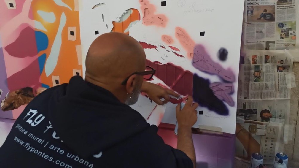 O artista Fly Pontes a pintar uma das camadas de stencil de um dos pardais. Do outro pardal, várias camadas já foram aplicadas, e são visíveis as marcações da técnica de stencil multicamadas. Foco na criação dos pássaros.
