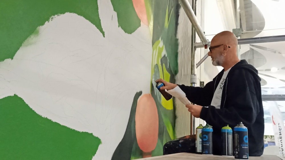 O artista Fly Pontes sentado no andaime, a pintar os verdes das folhas da laranjeira. Tem uma lata de spray numa mão e uma folha de referência na outra. Em primeiro plano, latas de spray à base de água (water based) no chão.