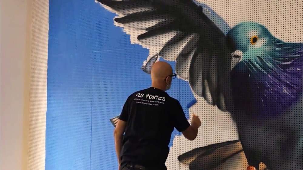 O artista Fly Pontes a pintar o fundo de azul com rolo, no mural "Tributo ao Pombo-Correio". Uma parte da cabeça e corpo do pombo principal já está visível, em contraste com o céu que está a ser preenchido.