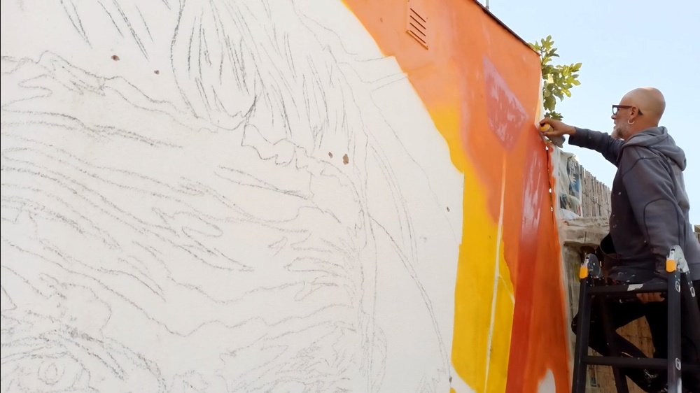 O artista Fly Pontes em cima do escadote a pintar o fundo de laranja com spray. Em primeiro plano, as linhas a carvão delineiam a cara de Nelson Francês, mostrando a fase de preparação do mural VEOH.