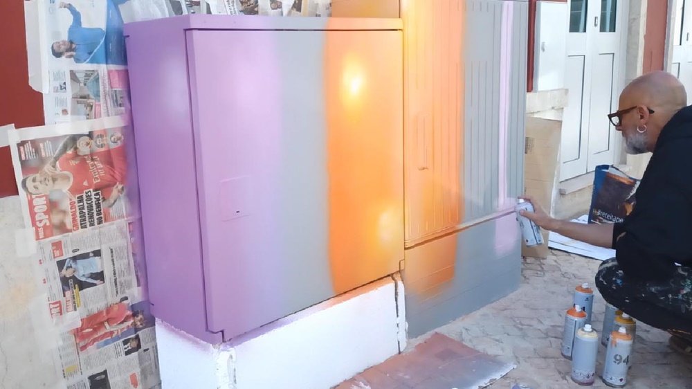 O artista Fly Pontes agachado a pintar o fundo do armário de electricidade. Já aplicou o violeta de um lado, o laranja ao centro, e está a pintar o rosa do outro lado. Partes do armário ainda mantêm a cor cinza original. Latas de spray visíveis.
