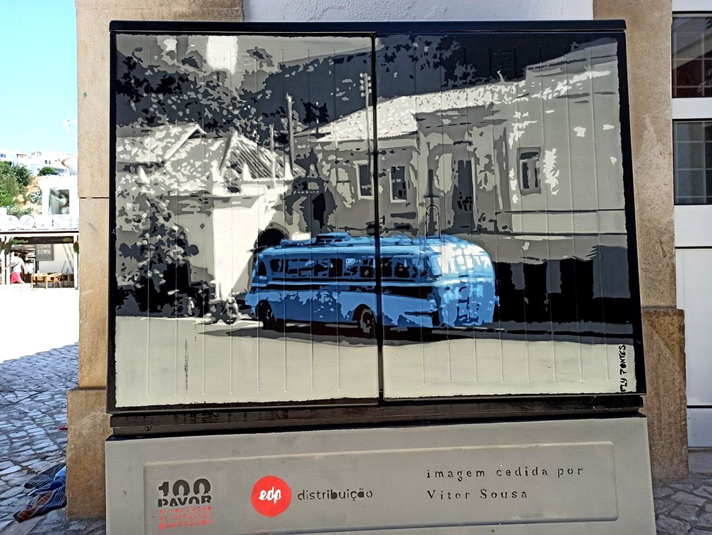 Pintura em Albufeira que recria a paragem de autocarro do Largo da Meia Laranja. O autocarro (Castelo & Caçorino) em azul contrasta com a arquitectura de fundo em tons de cinza, celebrando a memória urbana da cidade.