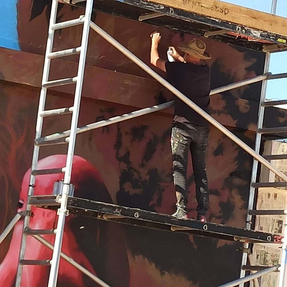 Artista Fly Pontes sobre o andaime, pintando detalhes de gorgónias e algas no mural Pedra do Valado. A parte superior do polvo é visível. Foco na flora marinha em arte urbana.