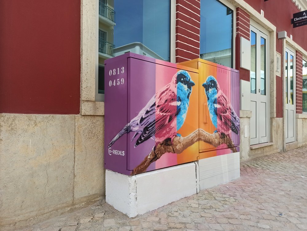 Vista lateral do armário de electricidade com a "Pintura dos Pardais" em Messines. É visível o prolongamento da cauda do pardal que se estende por este lado, unindo as diferentes faces da composição de Stencil e Spray.