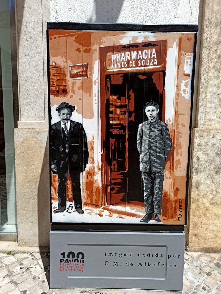 Pintura "A Primeira Farmácia de Albufeira" terminada num armário de electricidade. Os donos da farmácia estão em primeiro plano, pintados em tons de cinza (memória), contrastando com a fachada do edifício em tons quentes de castanho. Homenagem à história social de Albufeira.