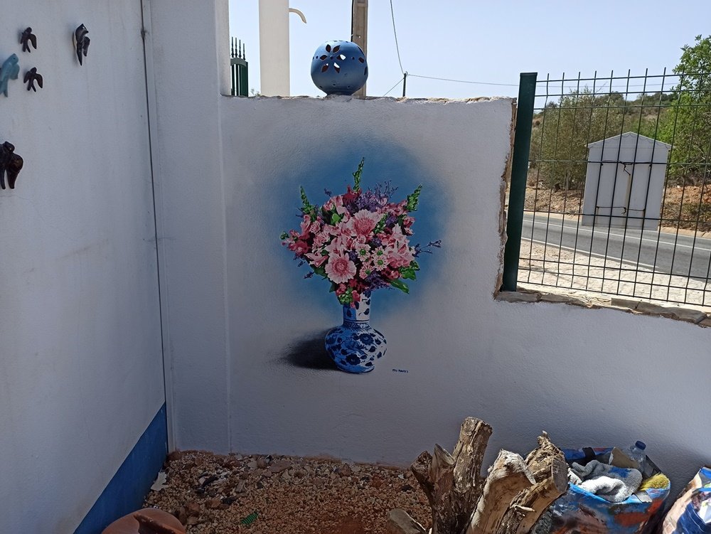 A pintura "Natureza Morta em Stencil" (bouquet e jarro) quase pronto, visto de uma perspectiva afastada. A estrada é visível no enquadramento, mostrando o contexto urbano da moradia particular.