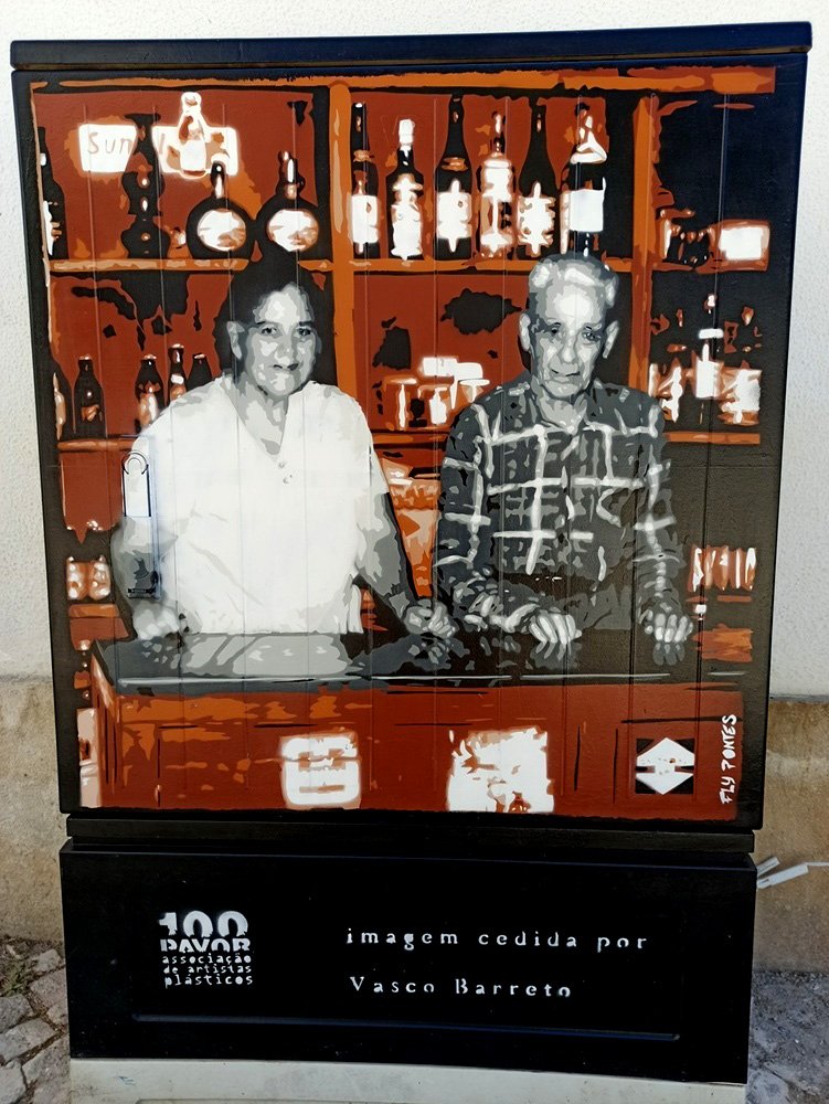Pintura "A Última Taberna de Albufeira" (stencil e spray) pintado num armário de electricidade. O casal de donos está representado em tons de cinza (nostalgia), contrastando com o ambiente da taberna (balcão, garrafas) em tons quentes de castanho.
