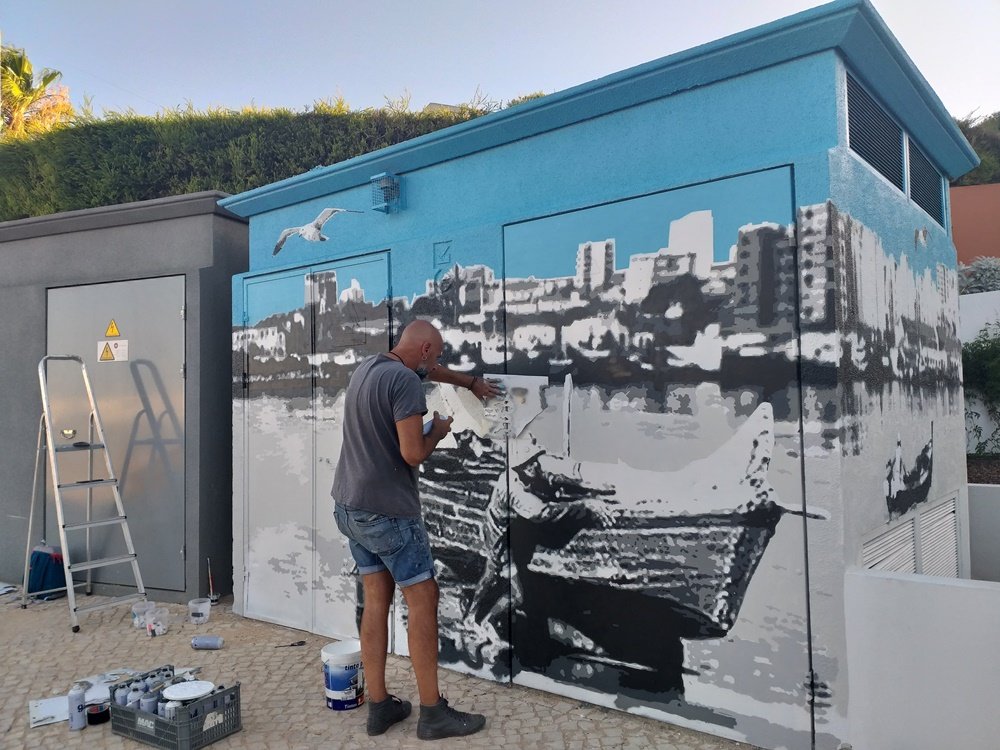 O artista Fly Pontes a pintar as cores do rio com um stencil na mão. A água é aplicada em vários tons de cinza, seguindo o esquema de cores do mural. As gaivotas e o céu azul contrastam com o resto da obra em tons de cinza.