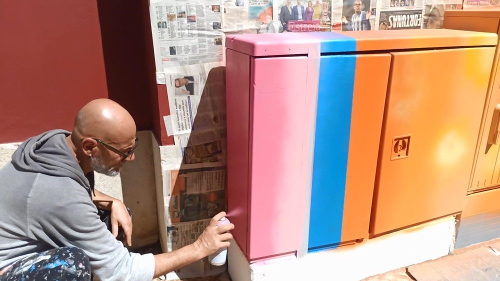 Processo de pintura do mural "Gatinhos Pop" em S. B. Messines. O artista Fly Pontes a pintar a lateral de rosa, enquanto a frente do armário de electricidade já tem o fundo laranja e azul aplicado.