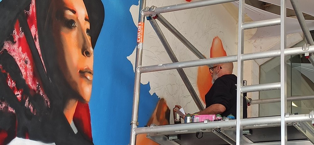 O artista Fly Pontes em cima do andaime, visto através dos seus ferros, a pintar o braço da jovem. Em primeiro plano, o rosto já tem os brilhos e os pormenores do lenço concluídos. Foco na pintura realista da pele no mural da Mostra da Laranja.