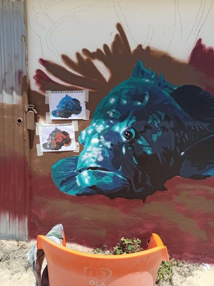 Pintura do Mero quase finalizada no Mural Pedra do Valado, em Armação de Pêra. Folhas de referência e a cadeira do artista no local.representando a fauna óssea do Recife do Algarve.