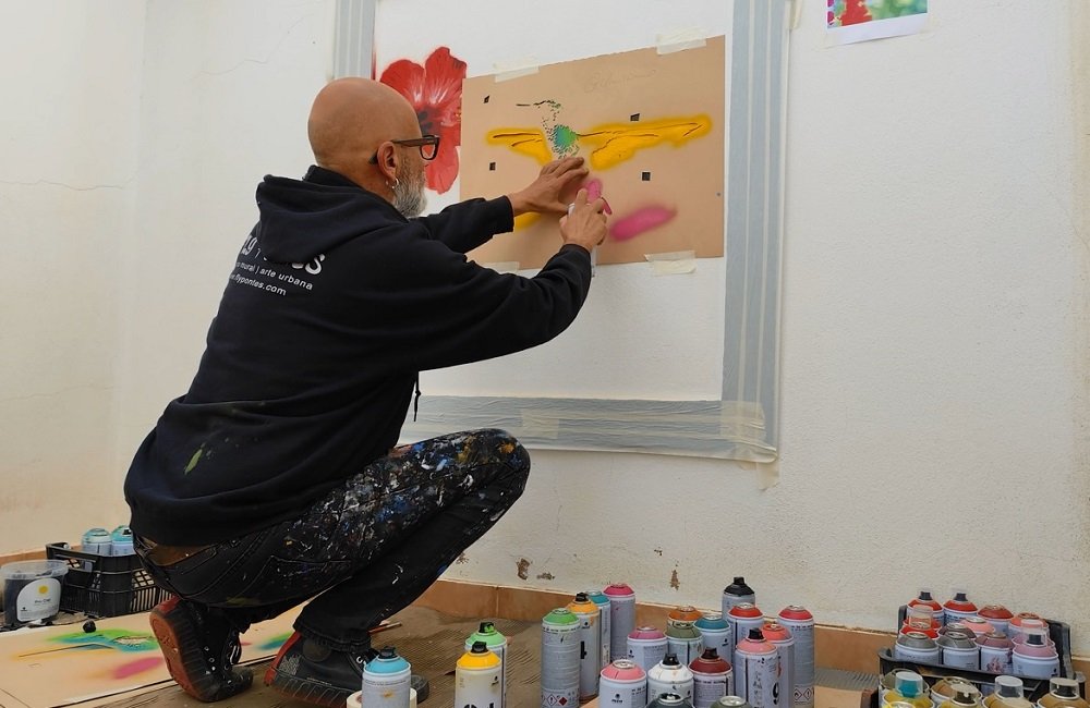 O artista Fly Pontes a pintar uma das camadas (layers) do stencil do Colibri, usando tinta spray. Vêem-se no chão as latas de tinta e as folhas das diferentes camadas do stencil. A pintura do Colibri na Janela Falsa em progresso.