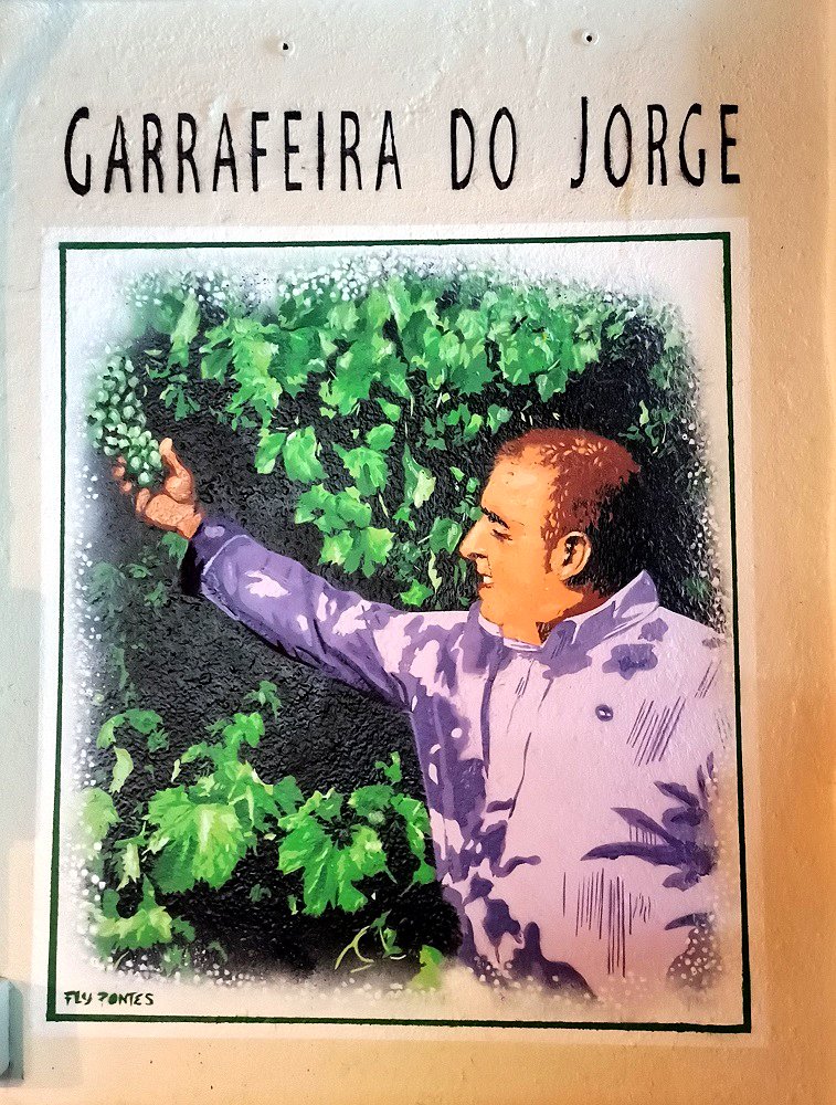 Mural na Garrafeira do Restaurante O Amândio em homenagem ao Jorge. Ele está em contemplação, a observar as uvas. A sua camisa violeta evoca os tons do vinho, simbolizando a sua paixão pela vinha.