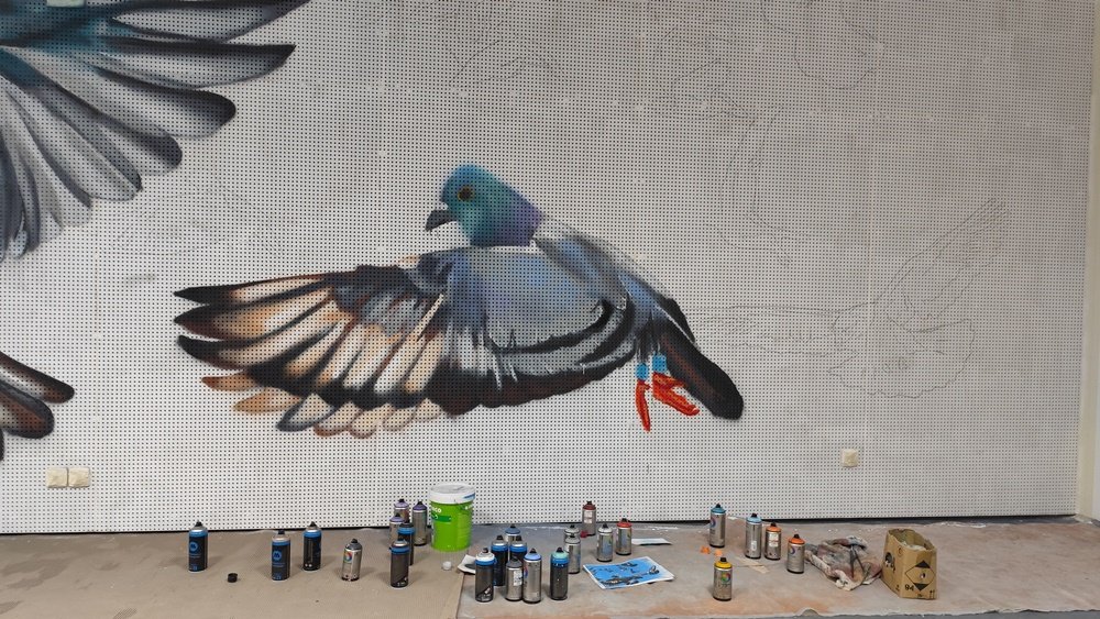 Pombo-correio de tamanho médio pronto no mural da Columbófila de Messines. As latas de spray usadas estão no chão e ainda se veem as linhas a carvão dos elementos por pintar, mostrando o processo de criação.
