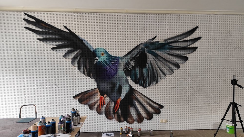 Pintura do pombo-correio principal (de asas abertas) quase pronta na Columbófila de Messines. Imagem ligeiramente afastada, mostrando latas de spray em cima da mesa e no chão, documentando o processo de criação de arte urbana.